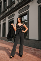
Beautiful brunette in black overalls, suit, posing on a city street, against the background of a new modern building in dark colors