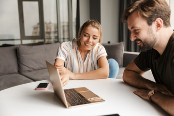 Photo of young joyful couple using laptop and smiling while sitting