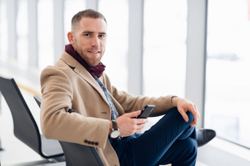 Attractive young adult man in stylish scarf and coat sitting inside airport terminal waiting area. He looking at cellular mobile phone screen and smiling