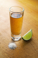 A tequila filled shot glass over a wooden tabletop with a slice of lemon  and a pinch of salt to the side.