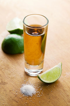 A tequila filled shot glass over a wooden tabletop with a slice of lemon  and a pinch of salt to the side.