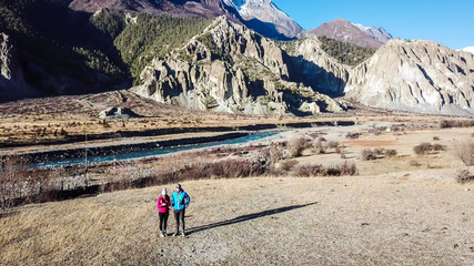 Obraz premium A couple walking on gravelled road, along the Annapurna Circuit Trek, Himalayas, Nepal. Snow caped Annapurna chain in the back. Clear weather, dry grass, snowy peaks. High altitude, massive mountains.