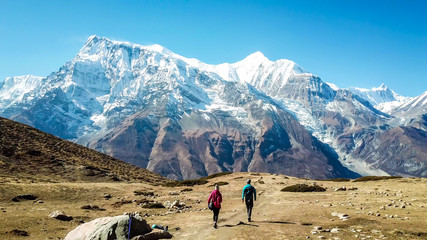 A couple walking on the Annapurna Circuit Trek, Himalayas, Nepal. Snow caped Annapurna chain in the back. Clear weather, dry grass, snowy peaks. High altitude, massive mountains. Freedom and adventure