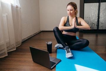 A sporty woman in sportswear is sitting on the floor with dumbbells and a protein shake or a bottle of water and is using a laptop at home in the living room. Sport and recreation concept.