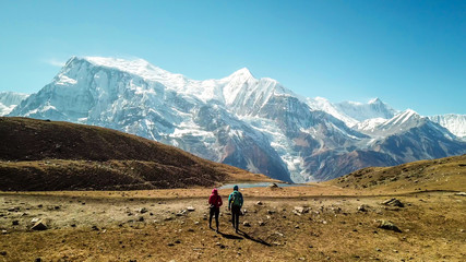 Fototapeta na wymiar A couple walking on the Annapurna Circuit Trek, Himalayas, Nepal. Snow caped Annapurna chain in the back. Clear weather, dry grass, snowy peaks. High altitude, massive mountains. Freedom and adventure