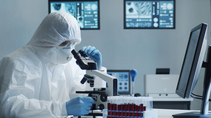 Scientist in protection suit and masks working in research lab using laboratory equipment: microscopes, test tubes. Coronavirus 2019-ncov hazard, pharmaceutical discovery, bacteriology and virology.