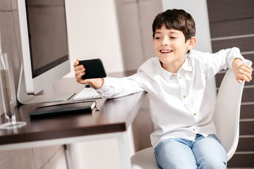 A boy standing in front of a computer taking a selfie with smartphone
