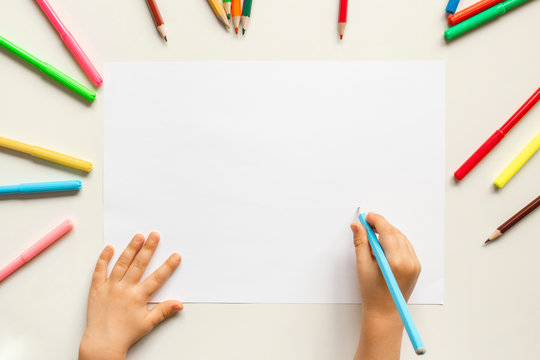 Top view of child's hands drawing at blank white paper within colorful pencils