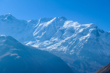 Fototapeta na wymiar A close up view on snow caped Himalayan peak seen from Annapurna Circuit Trek, Nepal. Sharp and steep slopes of the mountain. Powder snow being blown by strong wind. First sunbeams reaching the peak