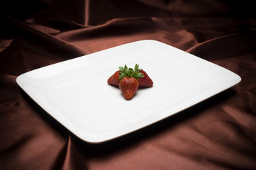 A small batch of strawberries in the center of a large white platter over an elegant low key cloth background.
