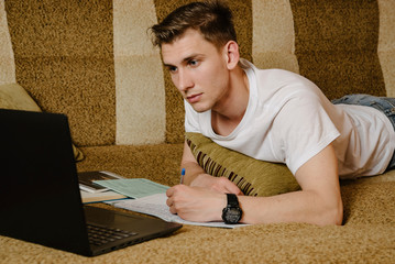 A young male student lying on a couch and using laptop while doing homework.Distance...
