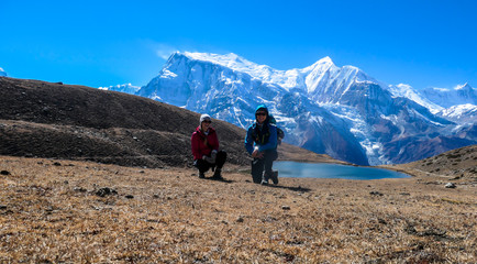 A couple in hiking outfits squatting next to a huge rock next to Ice Lake, Annapurna Circuit Trek, Himalayas, Nepal. High, snow caped Annapurna chain in the back. Happiness and love