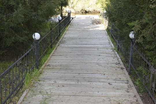 Wooden bridge with an iron fence and round glass lamps. Conifers over the fence along the entire length of the bridge. Exit to the river.