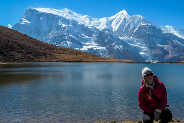 Fototapeta na wymiar A woman in hiking outfit squatting at the shore of Ice Lake, Annapurna Circuit Trek, Himalayas, Nepal. She is enjoying the idyllic landscape. High, snow caped Annapurna chain in the back. Achievement
