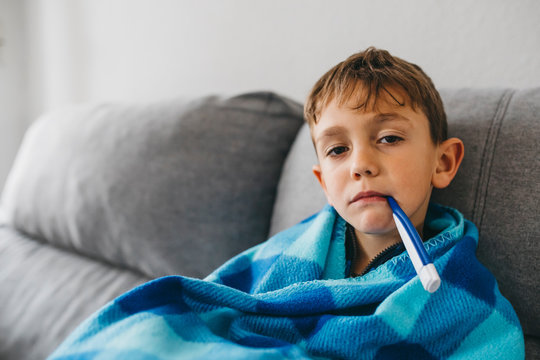 Portrait Of Sick Boy Sitting On Couch With Digital Thermometer In His Mouth