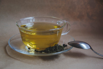Cup of green tea on brown background with dry leaves and small  jasmine flowers. Close up picture with fresh tea for bewering. Healthy drinks for break. Metal spoon on the side. 