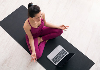 Stay home yoga. Young woman in lotus pose following online meditation on laptop, above view. Mockup for design