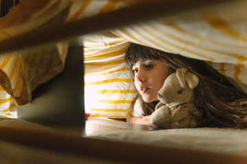 Portrait of girl with soft toy lying on bed under a blanket looking at laptop