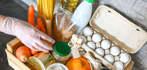 Volunteer hands in medical gloves holding donation box with various products for poor people. Pandemic coronavirus food shortages. Food donations or delivery concept. Free space for text copyspace