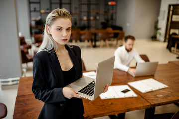 A young Manager girl standing in the office next to the workplace, with a laptop in her hands and looking directly at the camera