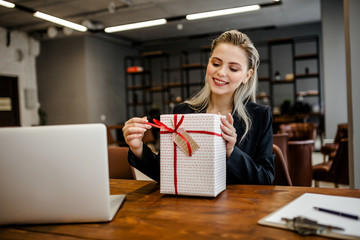 A beautiful blonde businesswoman sits at a Desk in the office and holds a gift that was given by colleagues at work