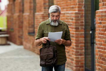Obraz premium A gray haired male businessman reads the details of a business contract standing on the street in front of the office