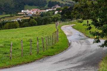 Fototapeta na wymiar Winding rural country road goes along fence with typical rural village and church of Basque Country, Spain. Camino de Santiago. Hiking and traveling concept.