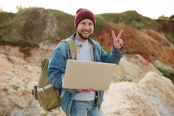 A bearded hipster man stands with a laptop in his hands, and shows a peace gesture