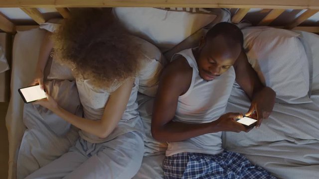Flat lay of African man wearing pajamas and his wife with curly hair lying back to back in bed and using their mobile phones