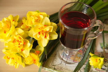 There is a glass cup of tea on the table, a notebook and a bouquet of beautiful yellow daffodils.