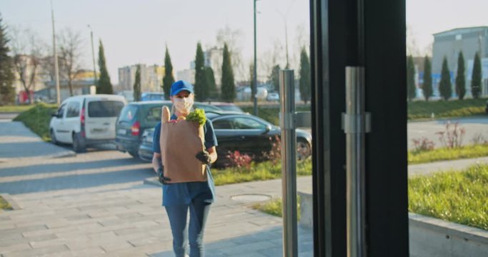 Rear of senior man coming out from building and meeting young Caucasian female courier from supermarket. Woman in medical mask and gloves handing packet with food to male. Pandemic concept.
