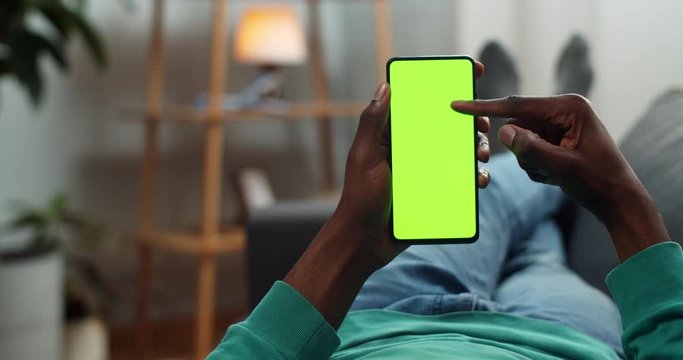 African american man scrolling mobile phone mock up screen while reading e book. Young man browsing net while lying on sofa. Concept of chroma key and green screen . Cosy home background.