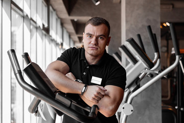 Fototapeta na wymiar Portrait of young handsome fitness trainer man in gym leaned on exercise bike while training around fitness equipment looking into the camera.