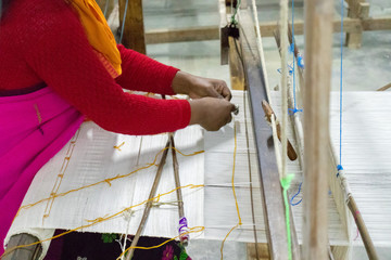 Lady demonstrating weaving of Mekhela (traditional Assamese dress) on the wooden hand-operated machine called loom using white & orange threads. A loom is a device used to weave cloth and tapestry. 