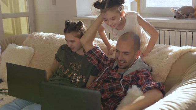 A Man With A Daughter-a Teenager Sitting On The Couch Playing On His Laptop. The Youngest Daughter Runs Up From Behind And Looking Curiously At The Monitor Screen.