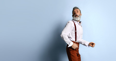 Elderly man in white shirt, brown pants and suspenders, bracelet. He runs looking back, posing sideways on blue background