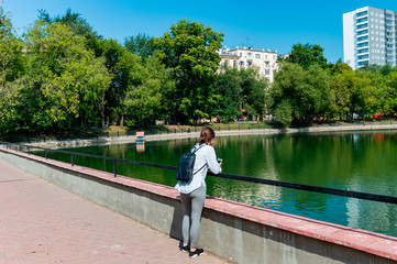 Fototapeta premium a girl stands leaning on the railing and looks at the still water of a pond on a Sunny summer day