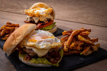 Homemade beef burger with fried egg, lettuce, a slice of tomato and french fries. Hamburger served on a rustic black table.