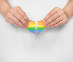 Human hands carefully glue together a broken paper heart painted in the colors of the LGBT flag: a...