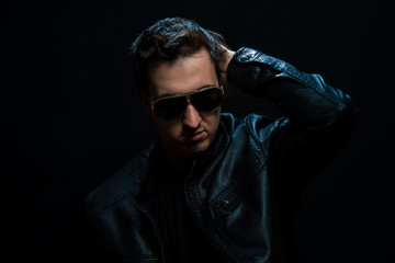 Obraz premium Young sexy stylish guy with glasses in a black leather jacket on a black background