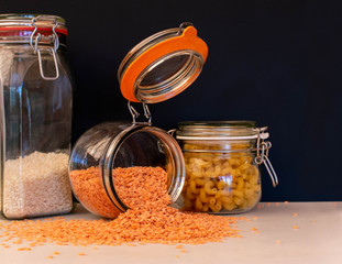Red lentils spilling out of a kilner glass storage jar with the lid wide open.  Storage jars containing dried food goods including rice and pasta sit either side against a plain black background