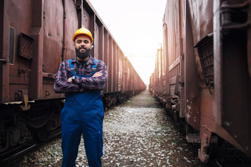 Portrait of railroad worker with crossed arms proudly standing at train station between wagons....