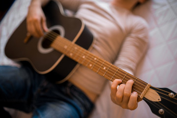 Young man in a light jumper and jeans plays an acoustic guitar in the morning in bed