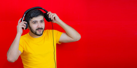 Handsome fashionable guy in a yellow shirt on a red background puts on headphones. Listening to music. Place for text.