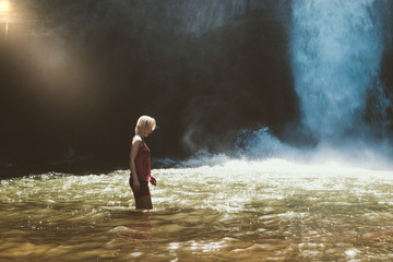 Obraz premium Young blonde woman with pink blouse and shorts walking in brown water river and illuminated by the sunbeam in front of the running blue waterfall in Bali island