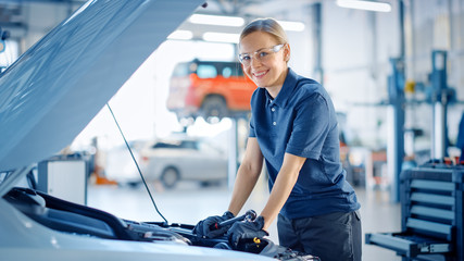 Beautiful Empowering Female Car Mechanic is Posing in a Car Service. She Wears Safety Glasses. Specialist Looks at a Camera and Smiles. - obrazy, fototapety, plakaty