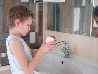 Happy boy washes hands with soap. Child playing with bar of soap and water in sunny bathroom with window. Water activities for children. Hygiene and skin care for children. Bathroom interior