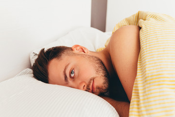 A young european man with beard is waking up in a bed on pillows and looking. Good morning