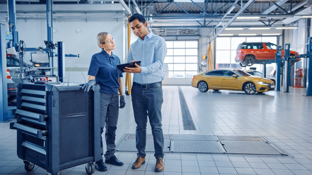 Car Service Manager And A Confident Female Mechanic Talk About Work Related Topics. They Use A Tablet Computer. Modern Clean Workshop With A Car On A Lift In The Background.