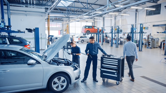 Two Mechanics in a Service are Inspecting a Car After They Got the Diagnostics Results. Female Specialist is Comparing the Data on a Tablet Computer. Repairman is Using a Ratchet to Repair the Faults.